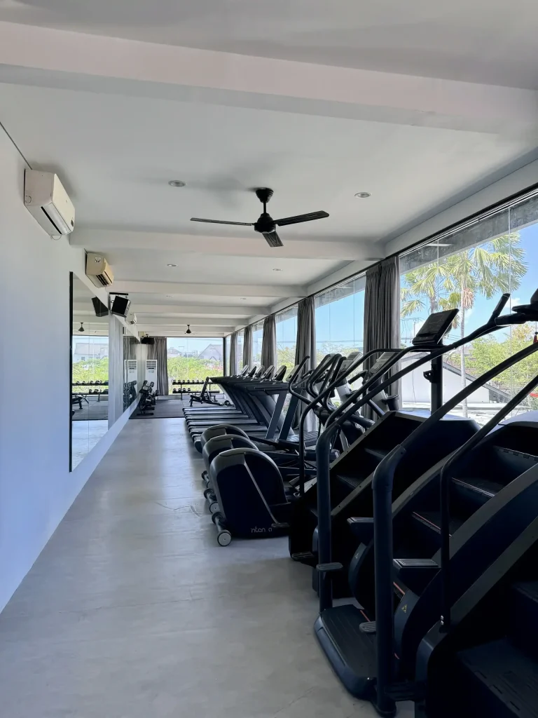 The cardio section on the second floor of Avenue Fitness Canggu, with a row of treadmills, stair climbers, and ellipticals next to large windows with palm tree views.