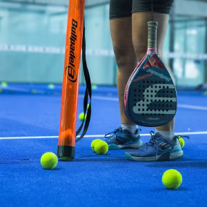 A close-up of padel equipment on a blue court at IndoPadel, including a Bullpadel racket, balls, and a ball collector.