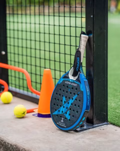 A Drop Shot padel racket leaning against the fence of a green padel court in Sanur, Bali, with balls and training cones nearby.