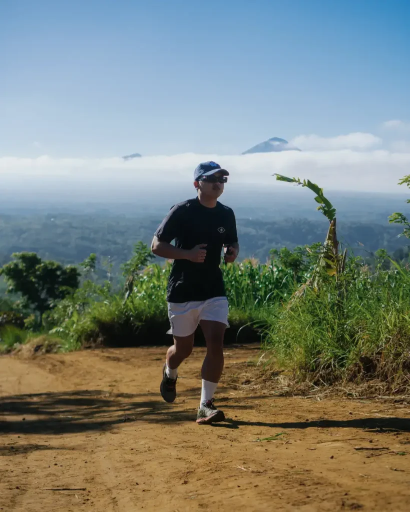 A participant in the Avenue Running Club Farm Trail Run event, running on a dirt trail with a scenic backdrop of Bali mountains above the clouds.